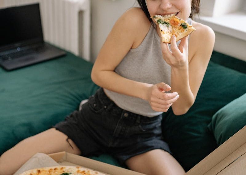 Woman in casual wear sitting on couch with pizza, looking happy and relaxed.