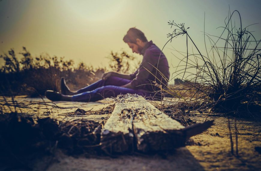 A man sits alone on sandy terrain at sunset, capturing solitude in nature's calm.