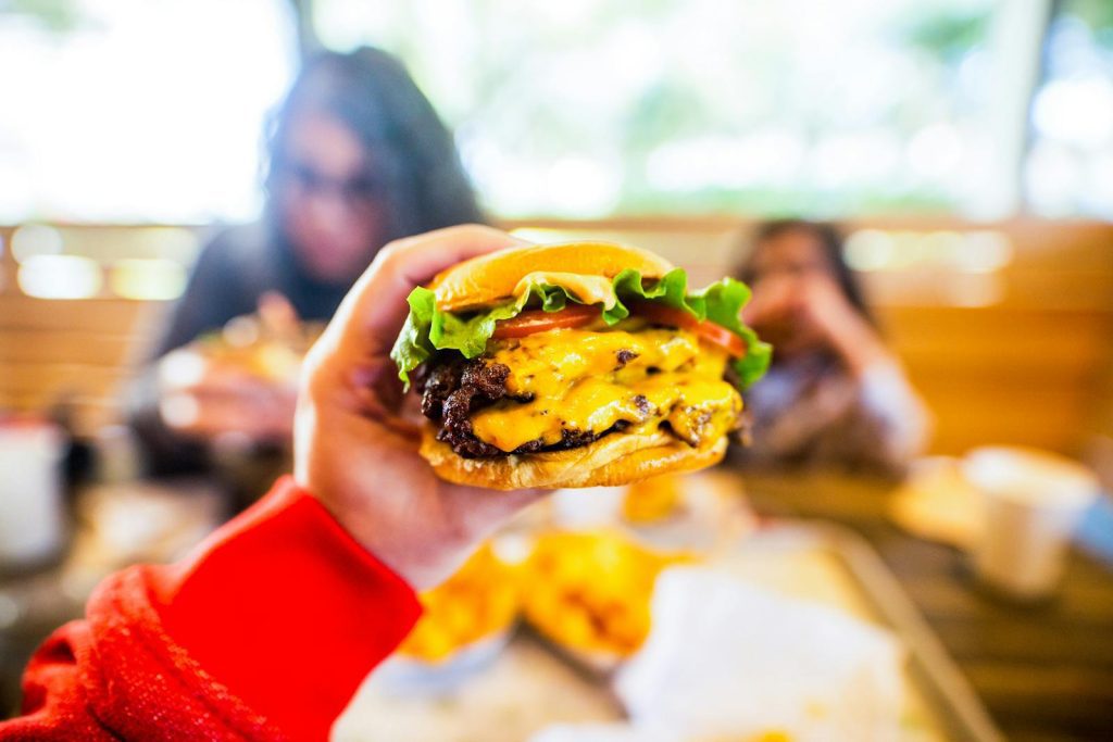 Crop anonymous person sitting at table and reaching out hand and demonstrating tasty cheeseburger in cafe in daylight