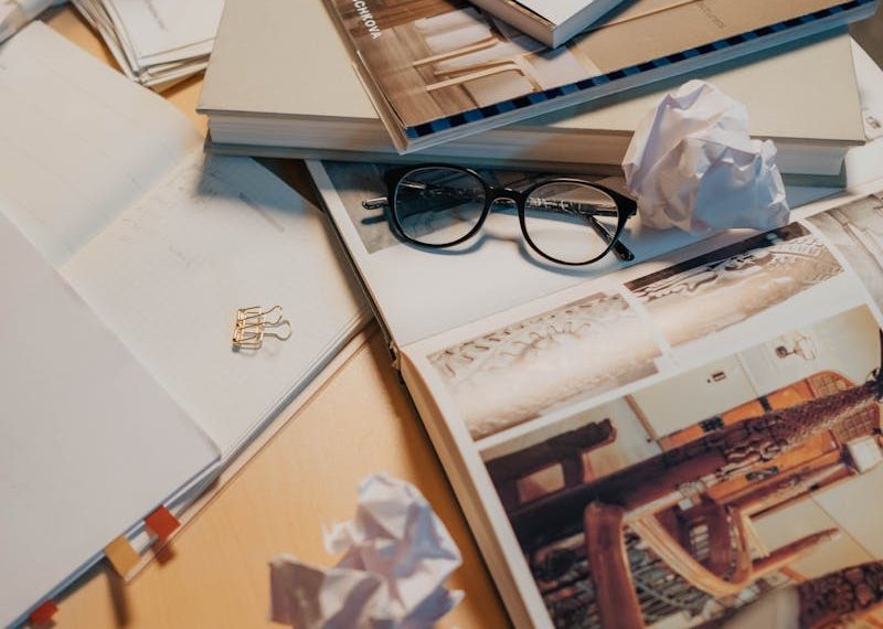 A cluttered desk with books, eyeglasses, crumpled papers, and a laptop, creating an unorganized workspace.