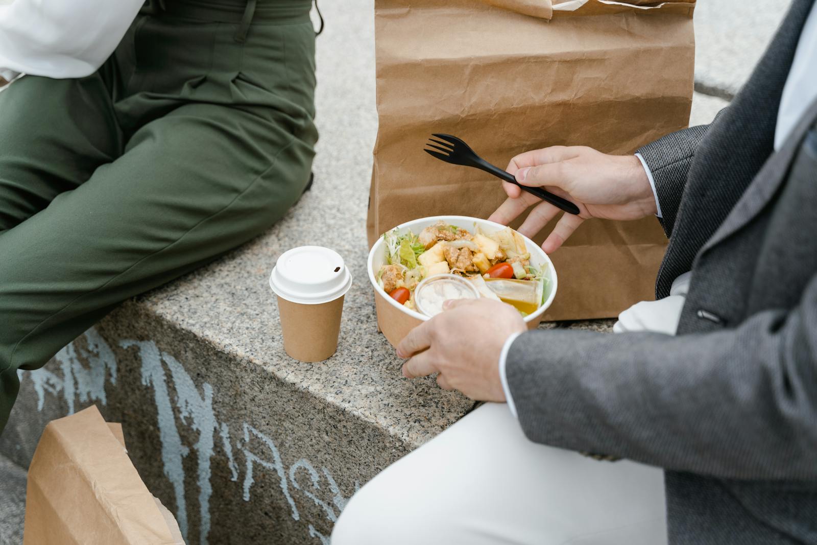 Two people enjoying a takeaway salad and coffee outdoors, seated on a stone bench.