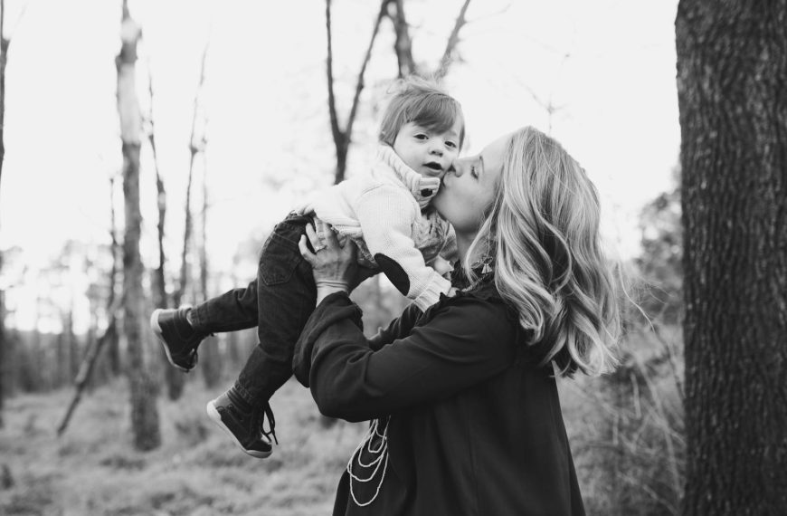 A touching moment of a mother kissing her young child in a serene outdoor setting captured in black and white.