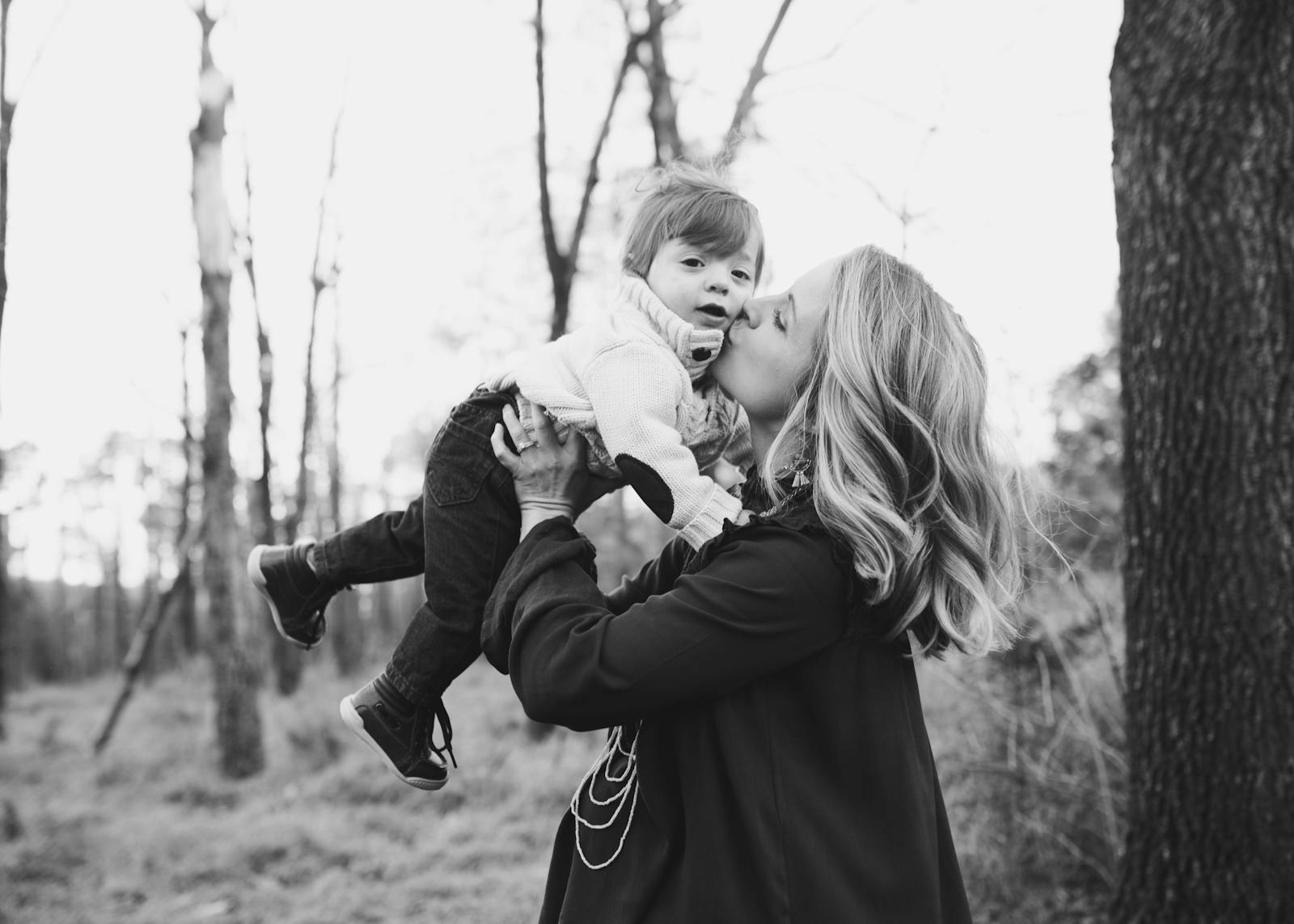 A touching moment of a mother kissing her young child in a serene outdoor setting captured in black and white.