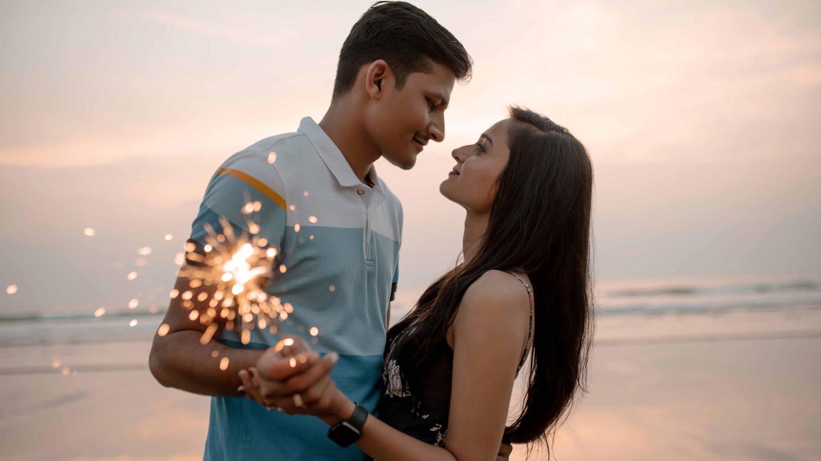 Loving couple holding sparklers at the beach during sunset, sharing a romantic moment.