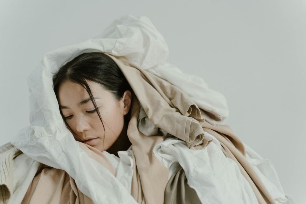 A serene woman enveloped in a pile of soft, neutral-colored clothes.