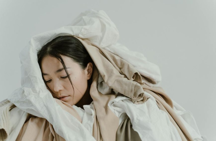A serene woman enveloped in a pile of soft, neutral-colored clothes.