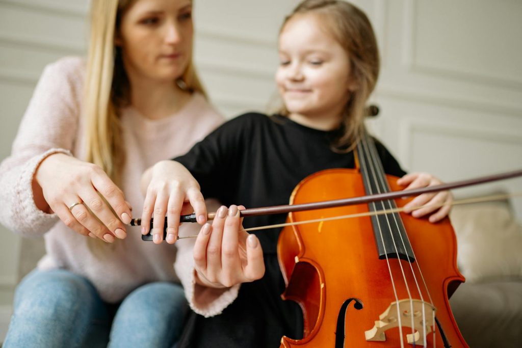A woman teaches a young girl to play the cello at home, focusing on positioning the bow.