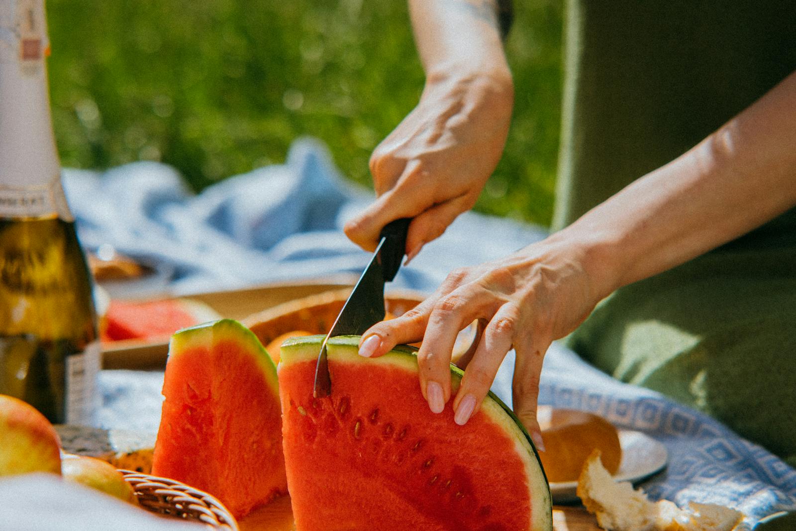 A close-up view of a person slicing watermelon during a summer picnic outdoors.