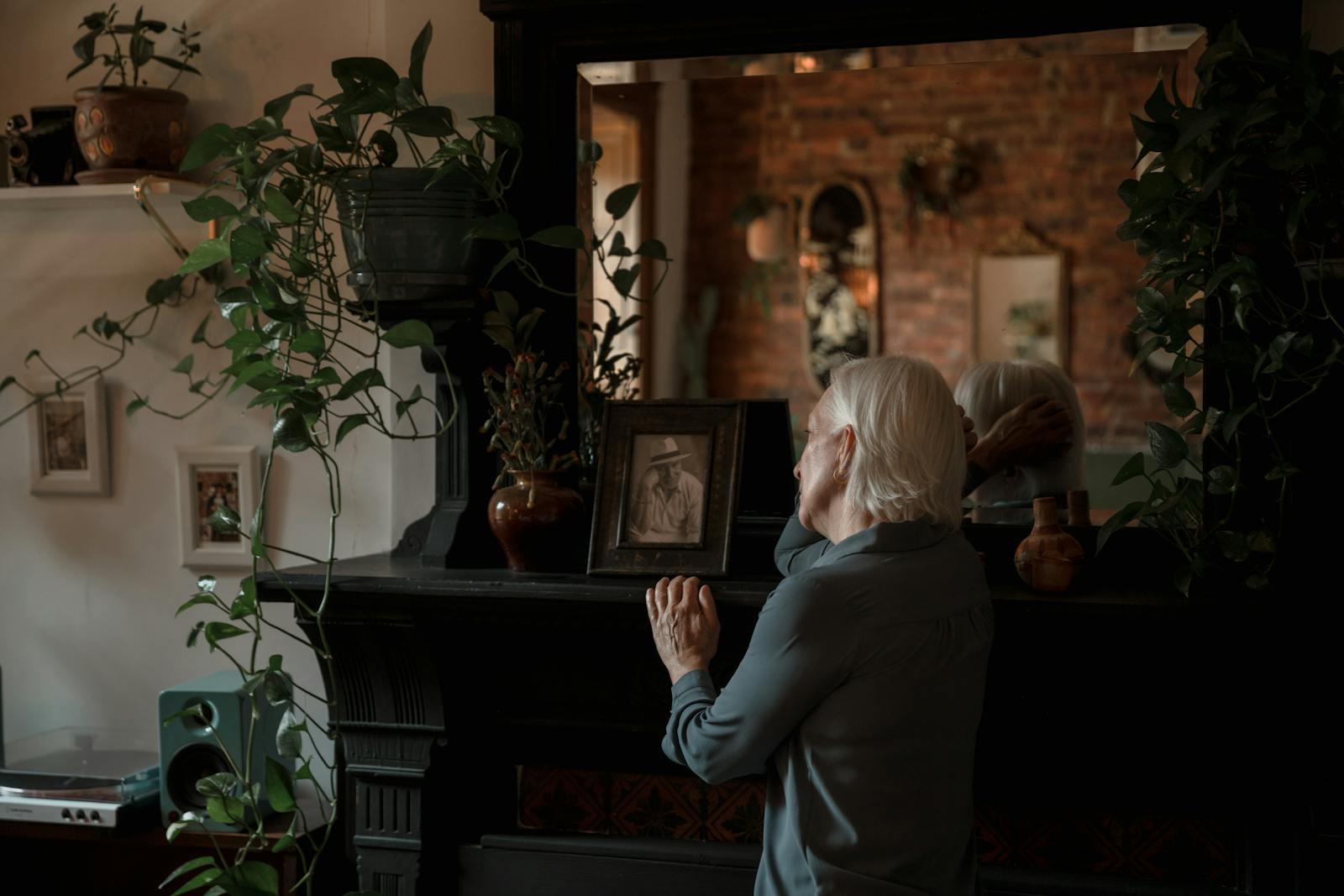 Senior woman reminiscing near a fireplace, surrounded by plants and picture frames in a cozy room.