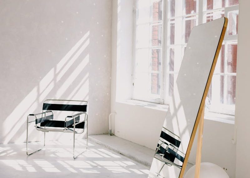 Bright minimalist room with a modern armchair, large mirror, and sunlight casting dramatic shadows.