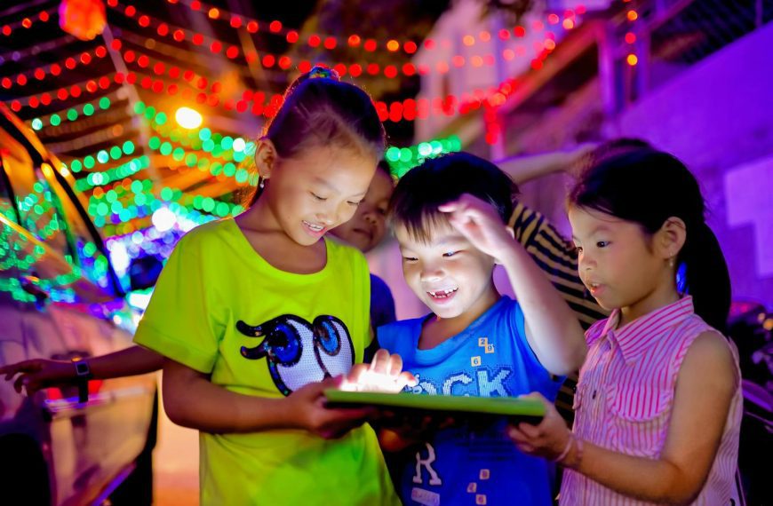 Group of happy children enjoying a night outdoors, playing together with a glowing tablet under colorful street decorations.
