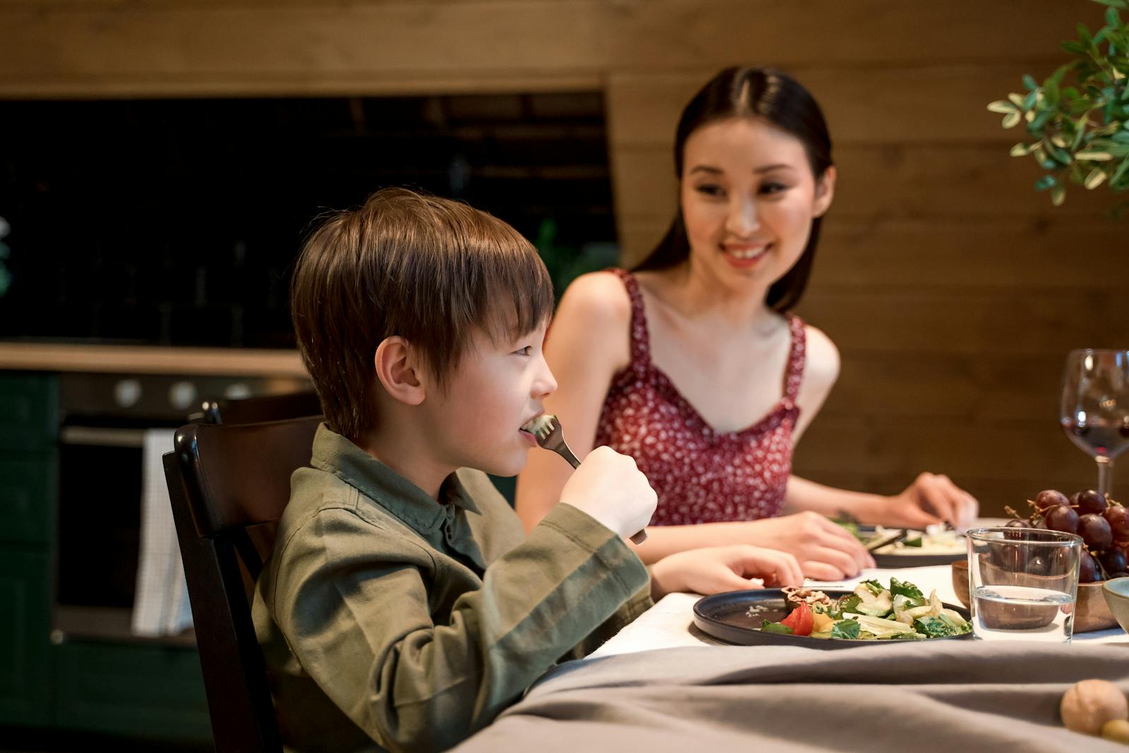 Asian mother and son dining indoors, happily eating a healthy meal with salad and vegetables.