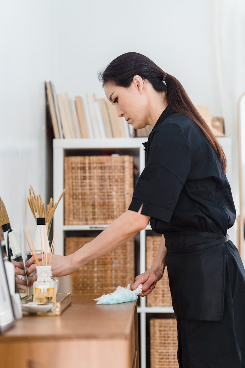 Woman in black uniform cleaning a wooden counter indoors with shelves in the background.