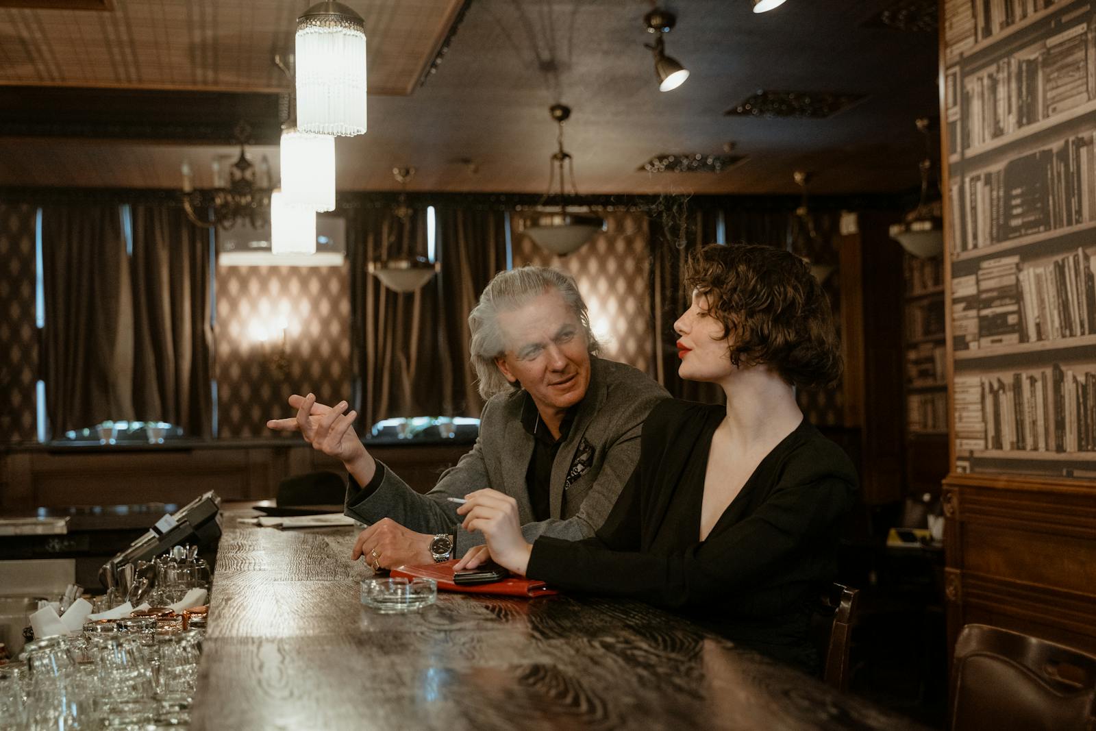 A senior man and stylish woman engaging in a conversation at an elegant, retro-styled bar.