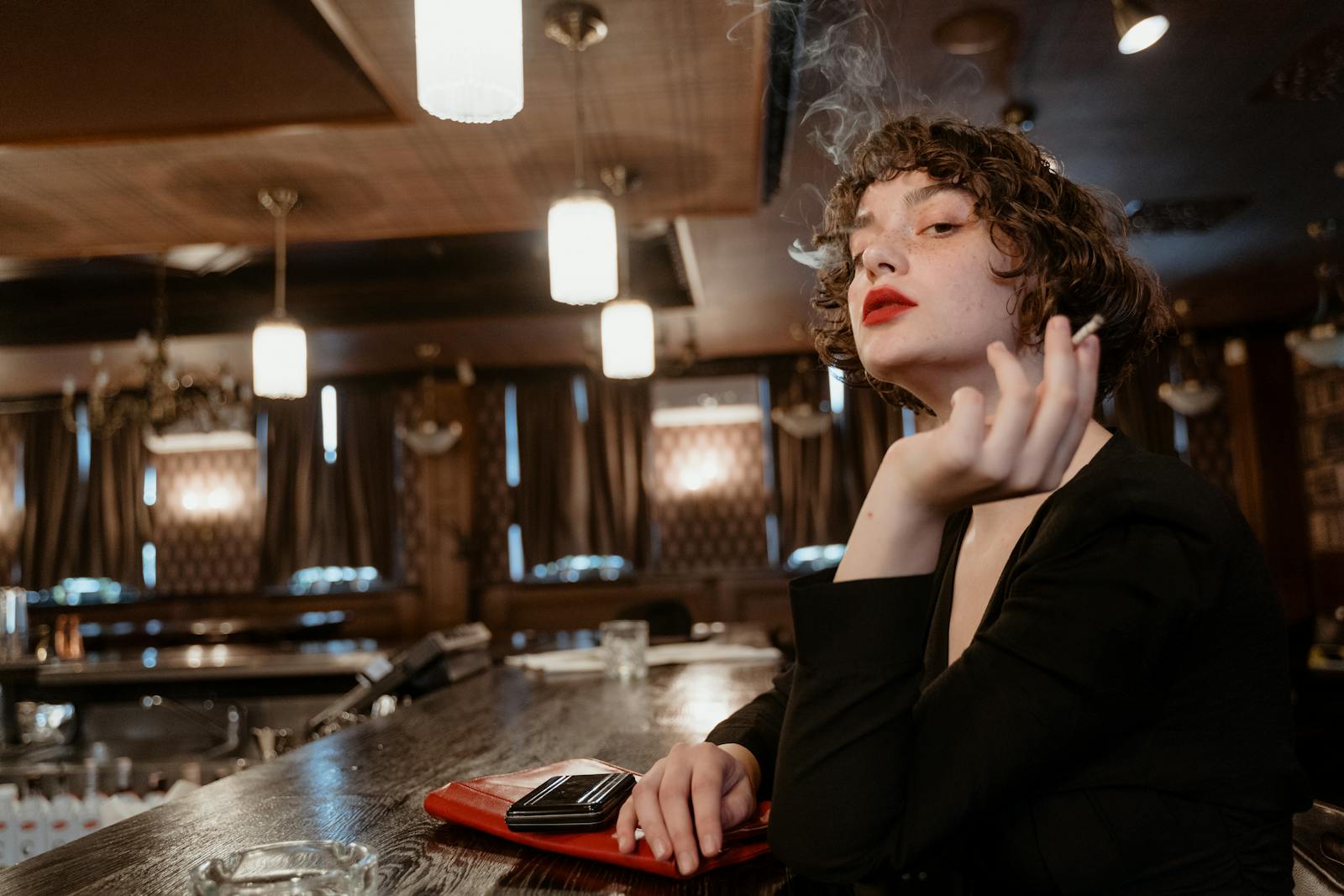 A fashionable woman with red lips sits smoking stylishly in a vintage bar.