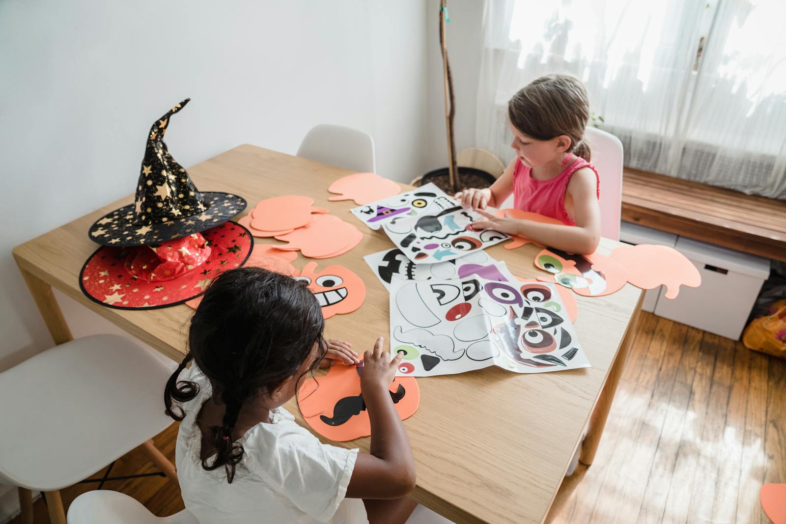 Two kids making Halloween crafts at a table with paper and witch hat.