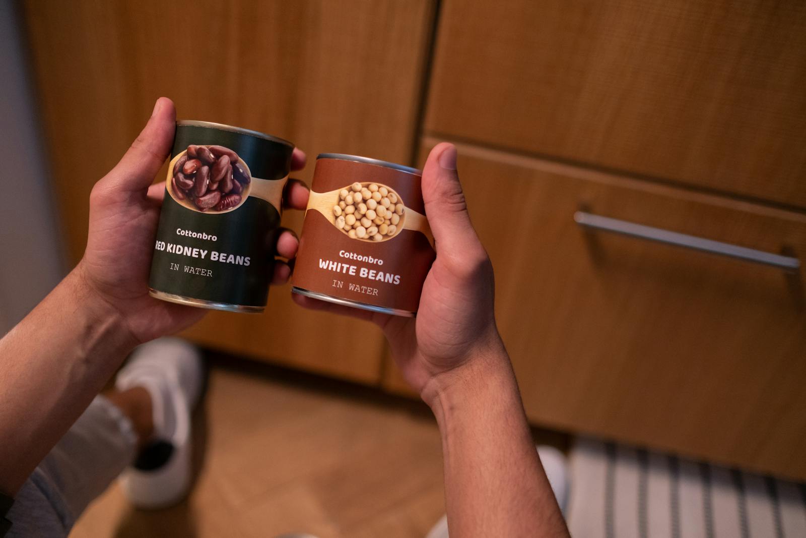 A person holding canned red kidney beans and white beans in a kitchen setting, emphasizing food storage.