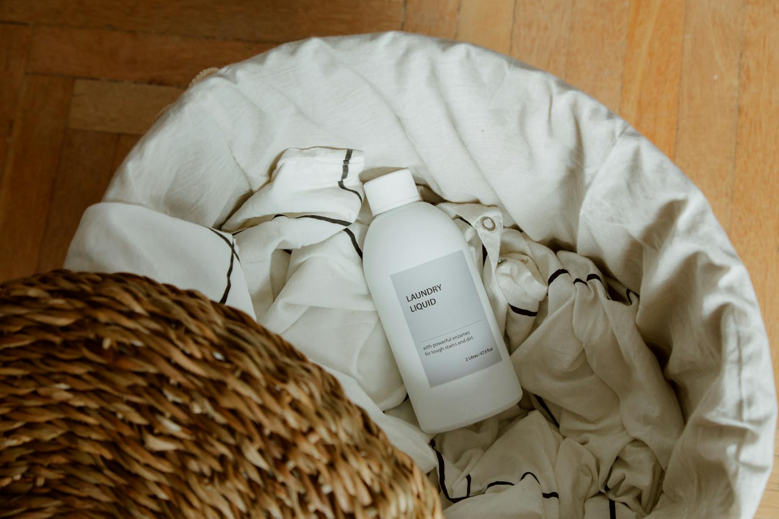 High angle view of laundry liquid in basket with white linens on wooden floor.