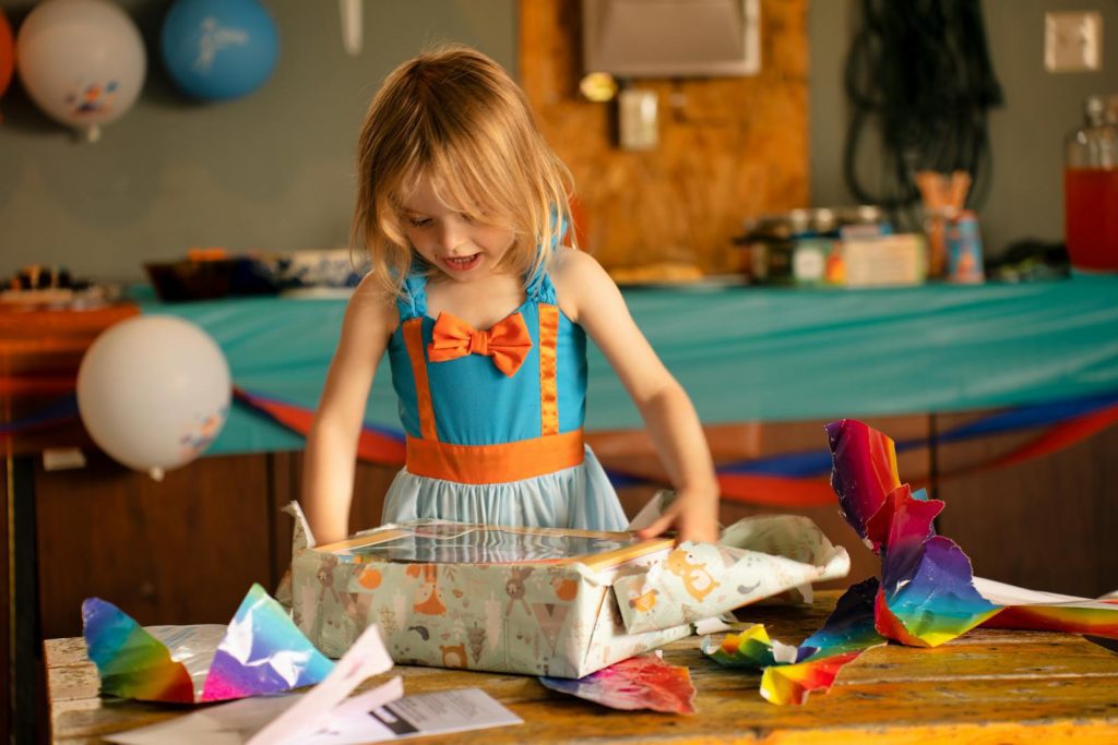 A happy girl in a blue dress opens a birthday present at an indoor party in Austin, MN.