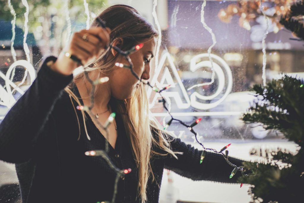 A woman decorates a Christmas tree with colorful lights indoors, creating a festive holiday ambiance.