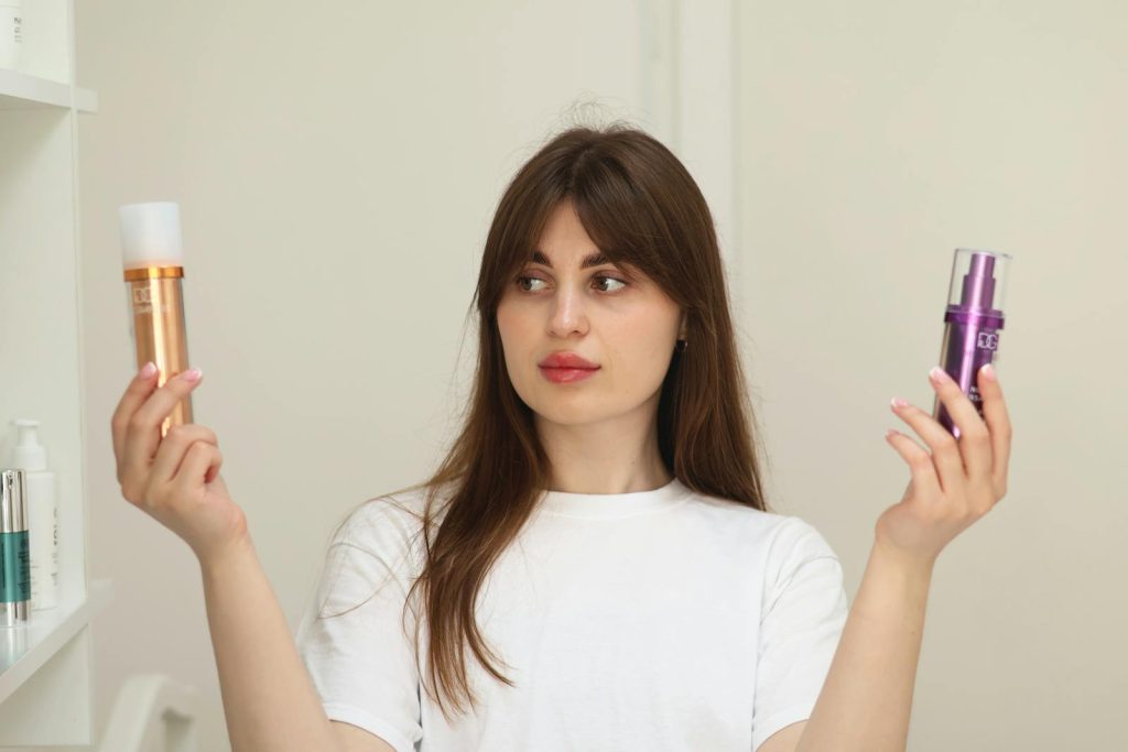 A young woman compares two cosmetic products while standing indoors, unsure of which to choose.