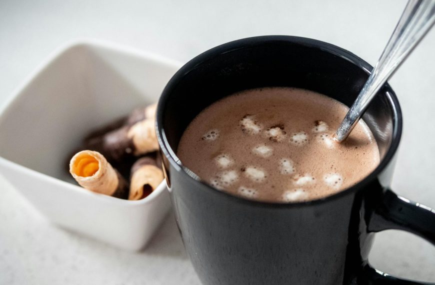 Delicious hot chocolate in a black mug with mini marshmallows and biscuit rolls on the side.