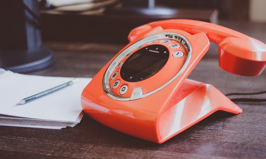 A retro-style orange rotary phone sits on a wooden desk alongside a pencil and notepad.