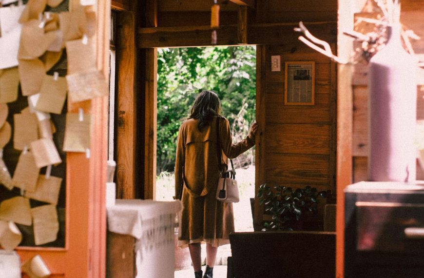 A woman in a warm coat steps out of a cozy cabin into the sunlight.
