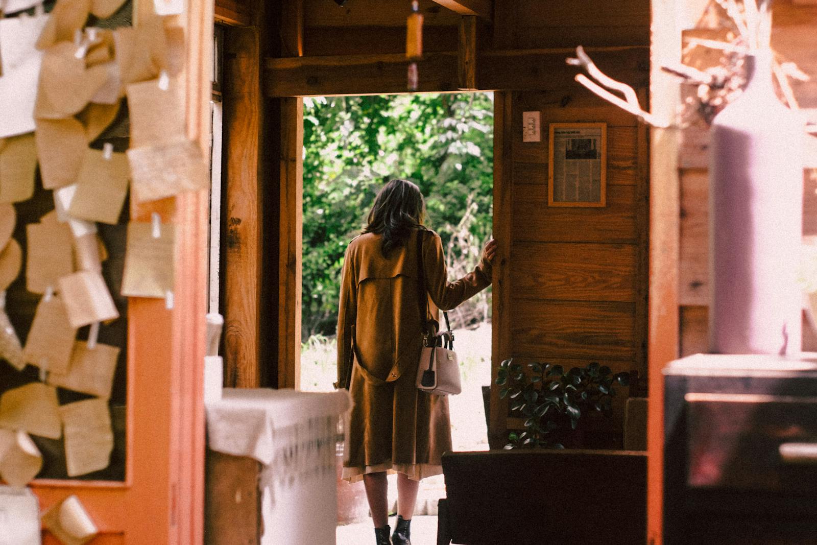 A woman in a warm coat steps out of a cozy cabin into the sunlight.
