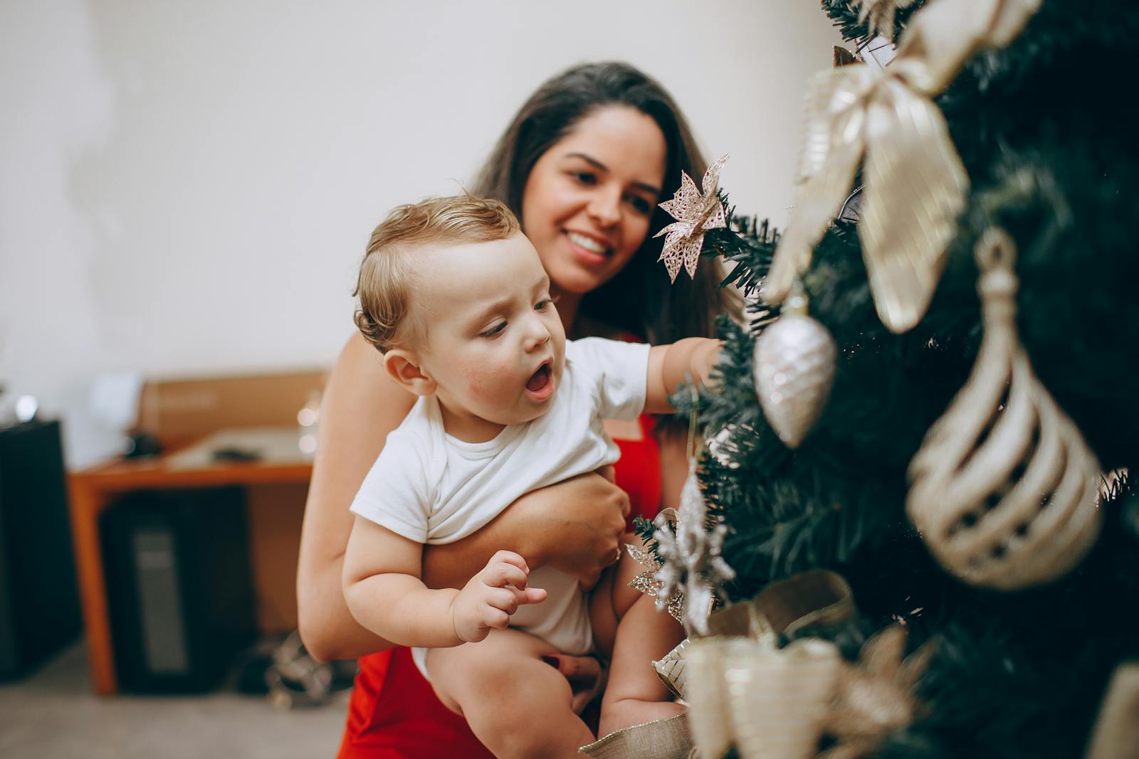 Smiling mother helps baby decorate a Christmas tree inside their home, capturing holiday joy.
