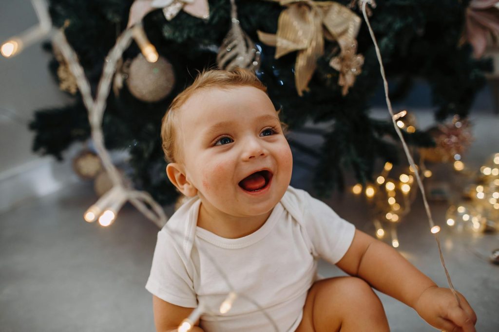 Adorable baby with Christmas lights, smiling by a decorated tree.