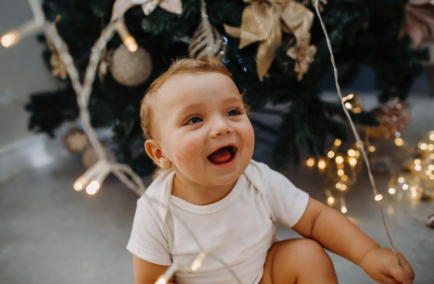 Adorable baby with Christmas lights, smiling by a decorated tree.