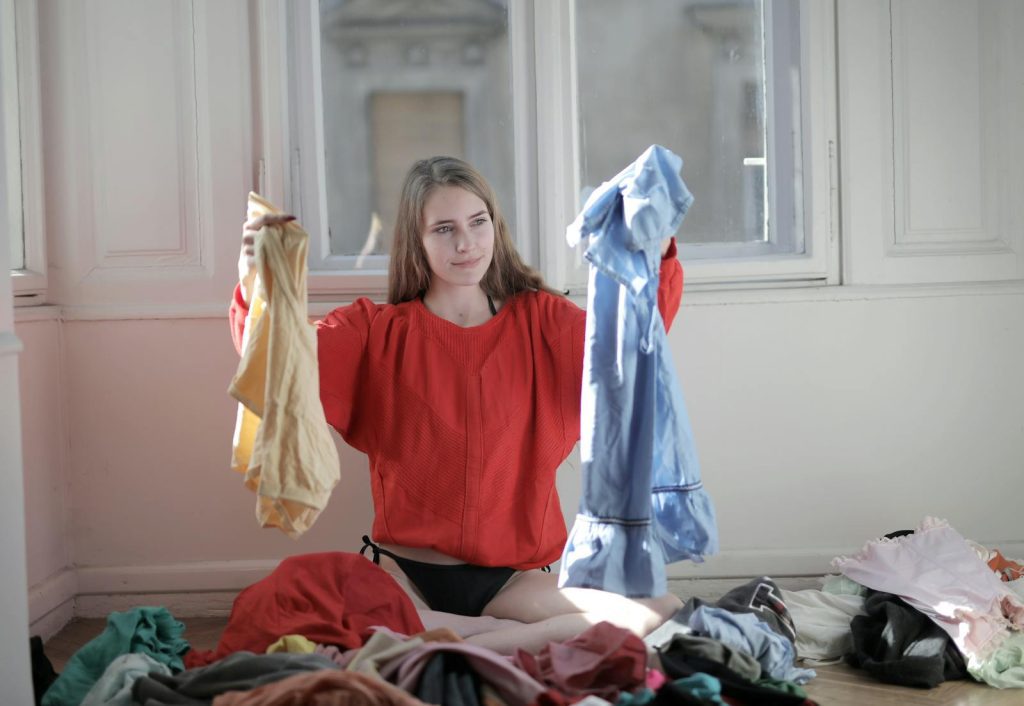 A woman in a red sweater deciding between clothes in a messy room.