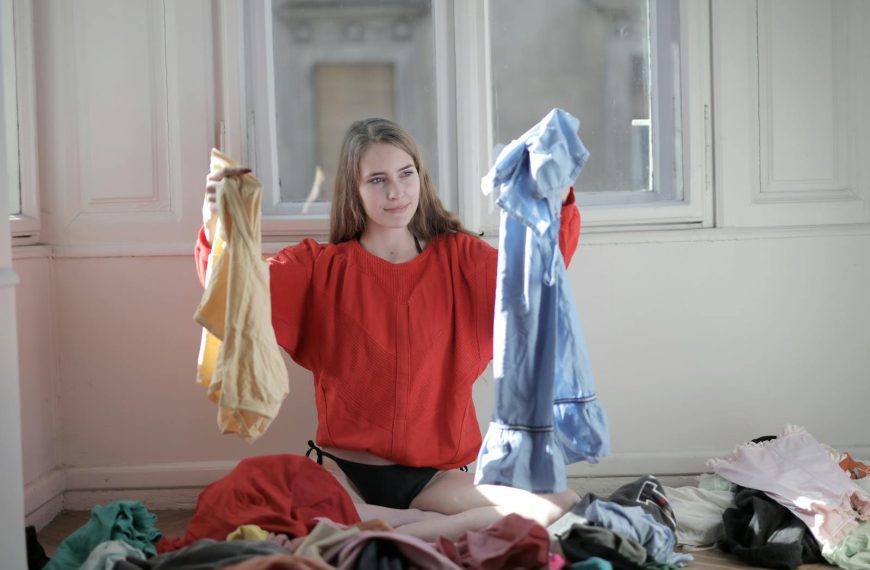 A woman in a red sweater deciding between clothes in a messy room.