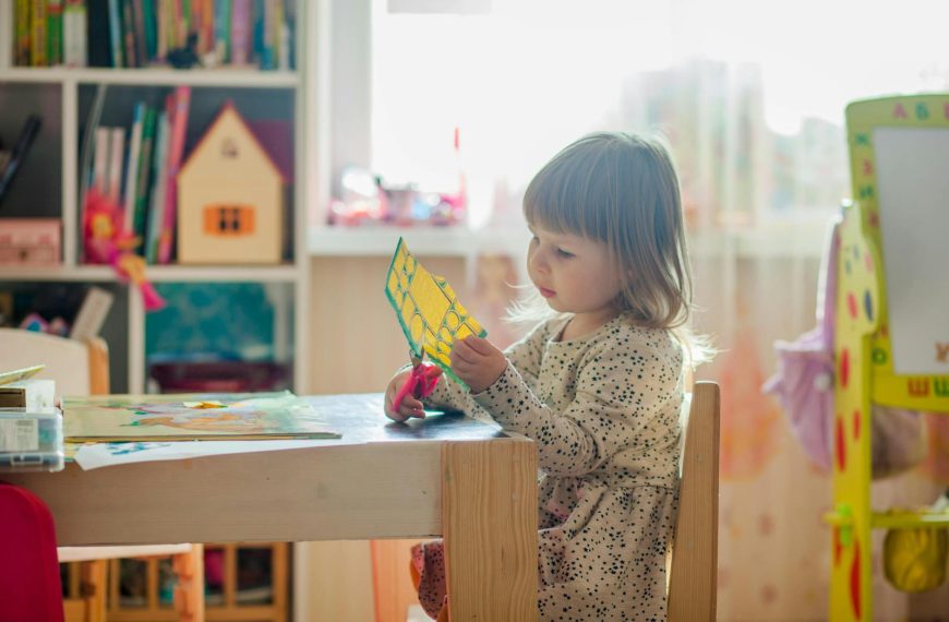 A young girl in a bright classroom engaged in a creative arts and crafts project at a table.