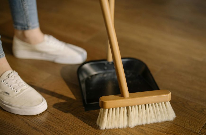 Close-up of cleaning process with broom and dustpan beside sneakers on a wooden indoor floor.
