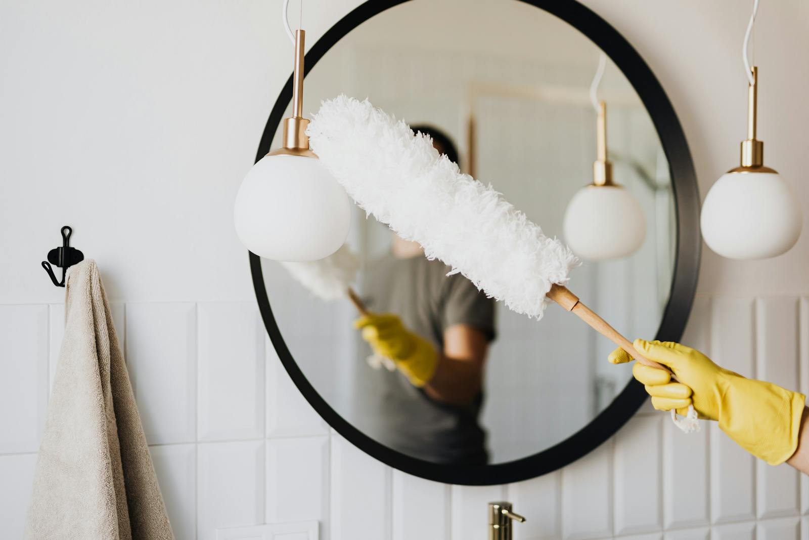 A person cleaning an elegant bathroom mirror with a fluffy duster and yellow gloves.