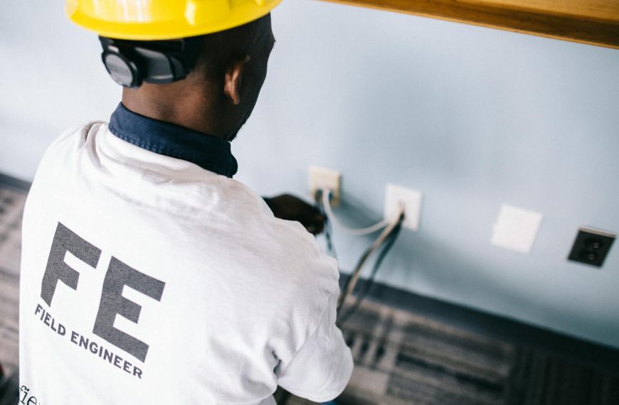 From above back view unrecognizable black field engineer wearing white shirt and protective hardhat sitting on floor and checking voltage in wall plugs