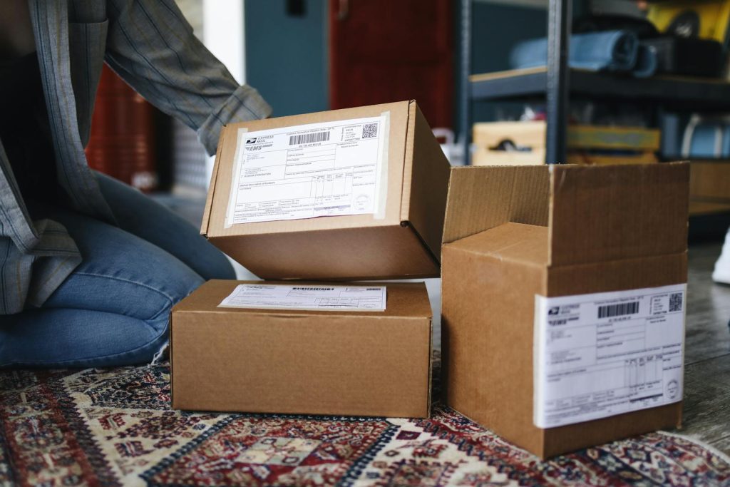 A woman kneels on a rug surrounded by cardboard packages in a home setting.