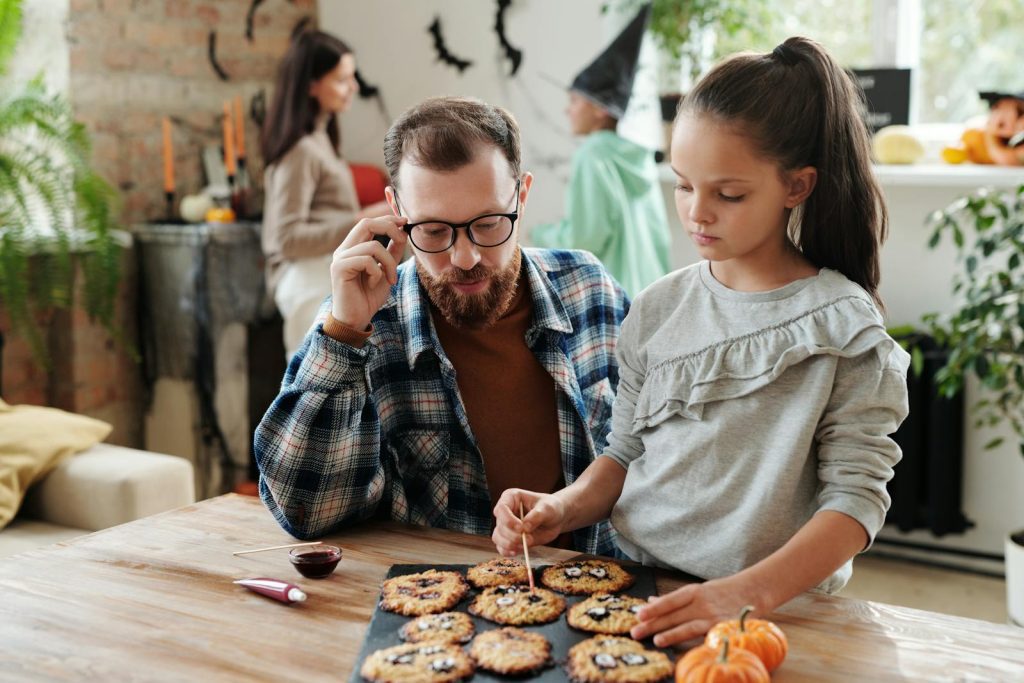 A father and daughter enjoy decorating Halloween cookies together in a cozy indoor setting.