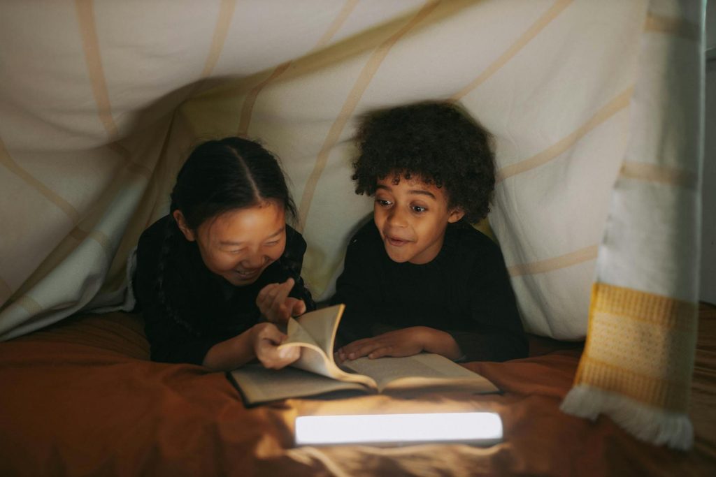 Two children read a book together under a cozy blanket fort, illuminated by soft lighting.