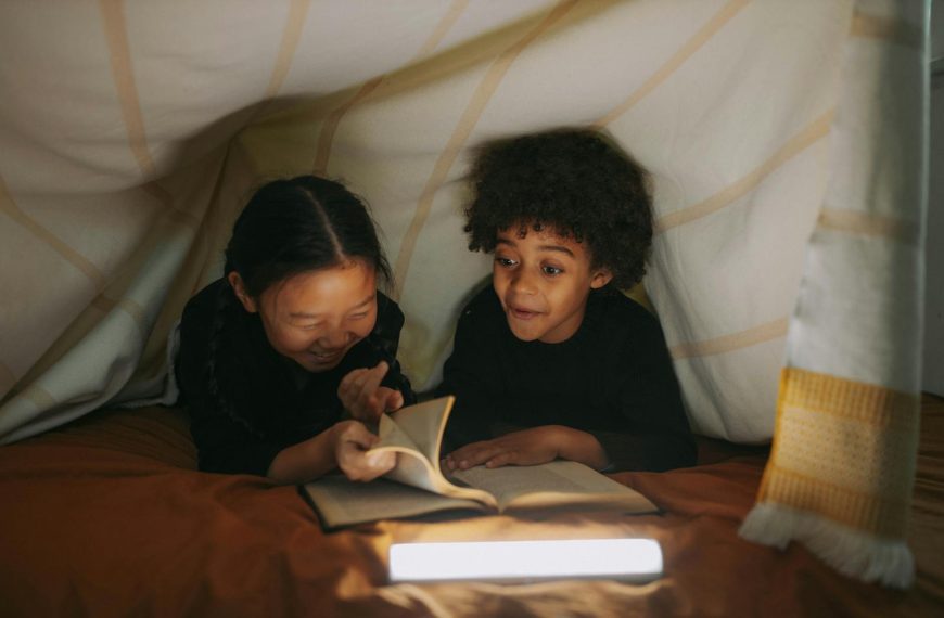 Two children read a book together under a cozy blanket fort, illuminated by soft lighting.