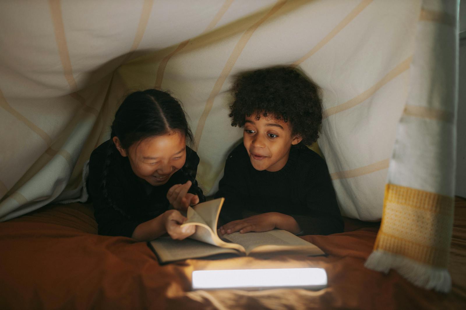 Two children read a book together under a cozy blanket fort, illuminated by soft lighting.