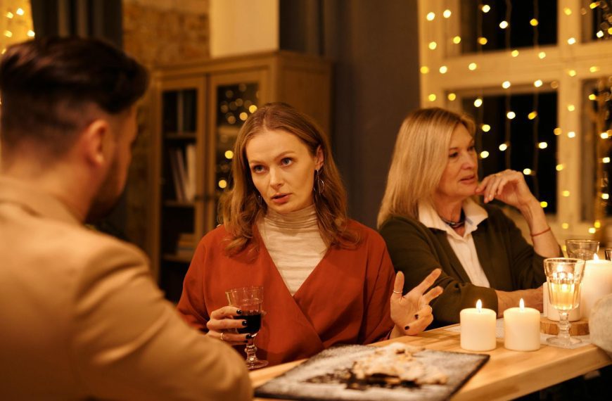 Three people enjoying a festive dinner table conversation surrounded by warm holiday lights.