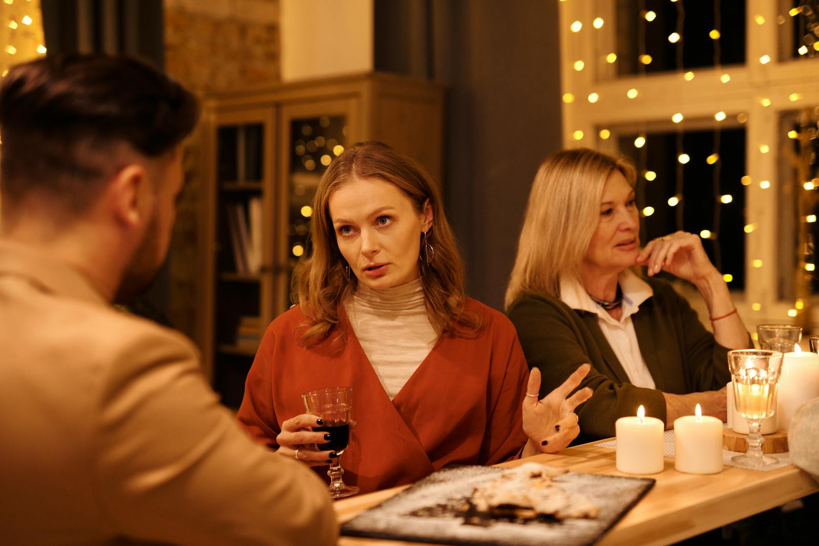 Three people enjoying a festive dinner table conversation surrounded by warm holiday lights.