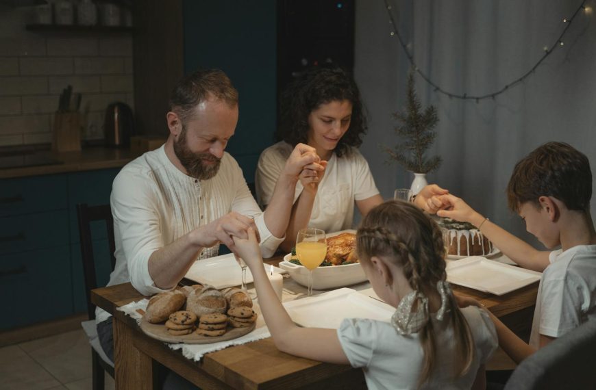 Family holding hands and praying over a dinner table with turkey and cookies during festival celebration.