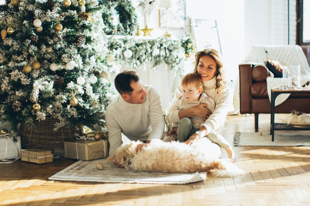 Family of three with a dog sitting by a decorated Christmas tree in a cozy living room.