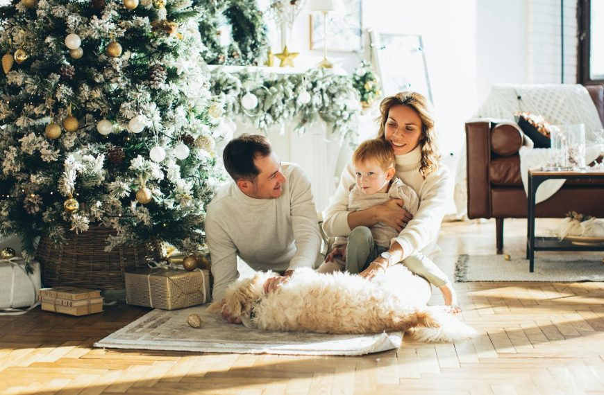 Family of three with a dog sitting by a decorated Christmas tree in a cozy living room.