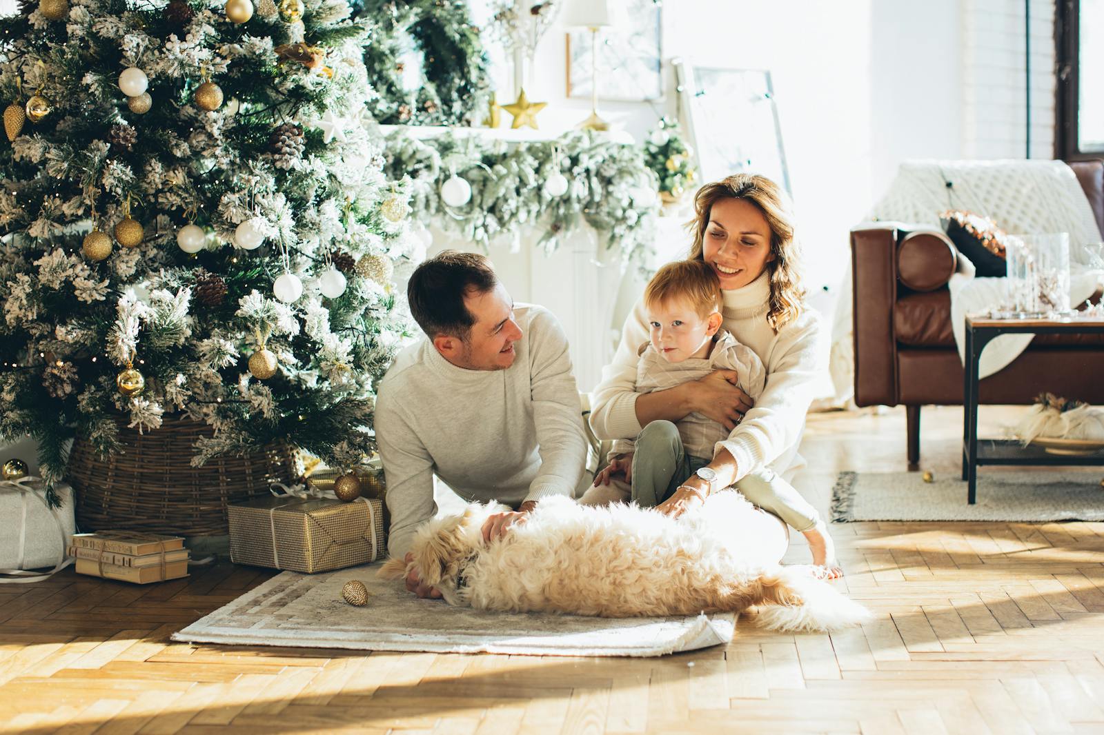 Family of three with a dog sitting by a decorated Christmas tree in a cozy living room.
