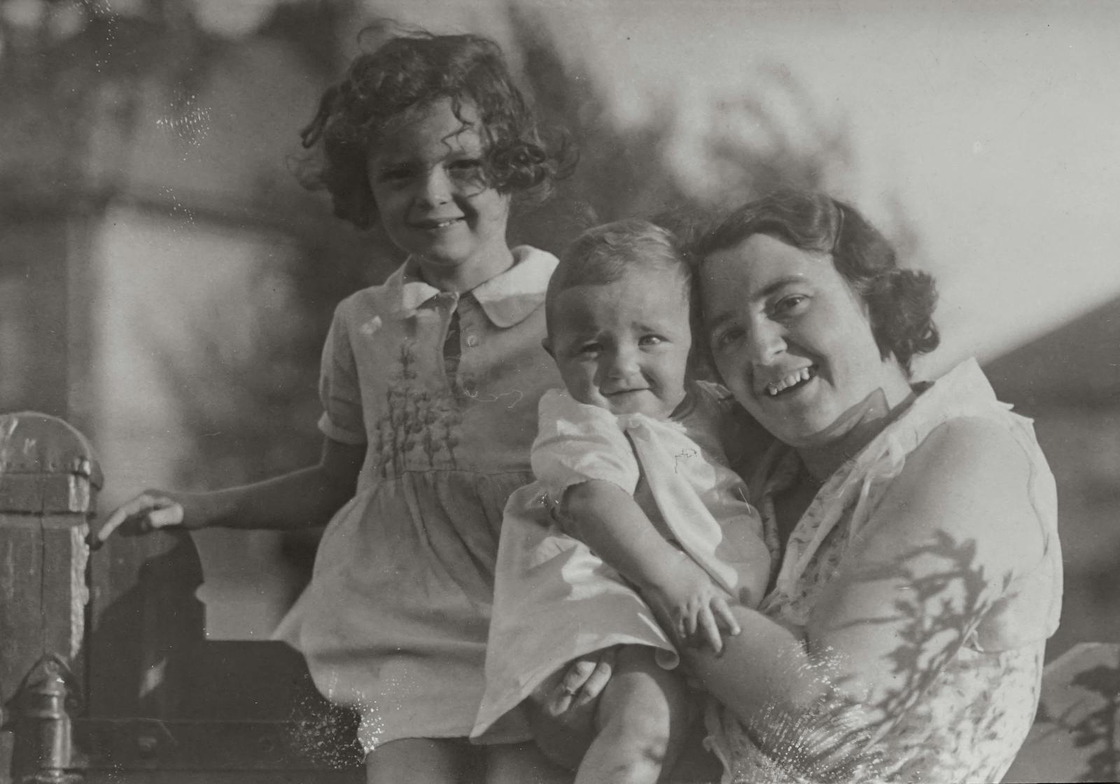 An old grayscale photo of a mother and her two children smiling outdoors.